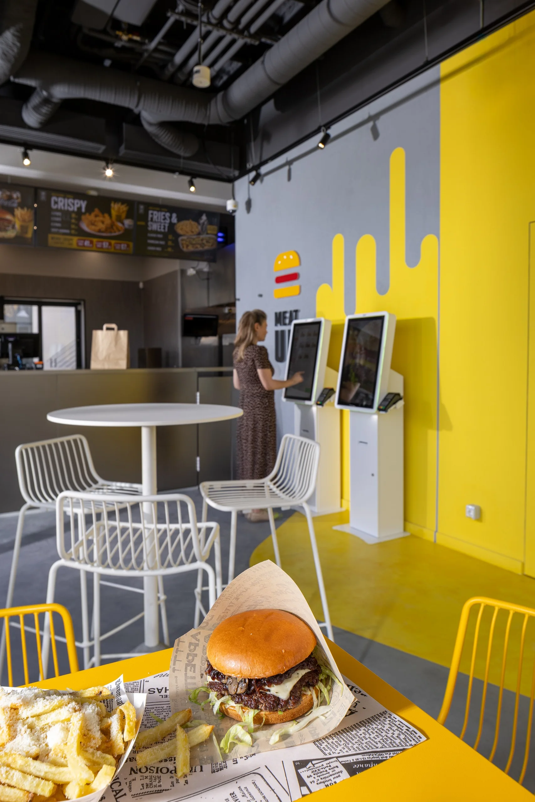 MeatUp - burger cu cartofi pe masa galbenă, cu sala și chioșcurile în spate